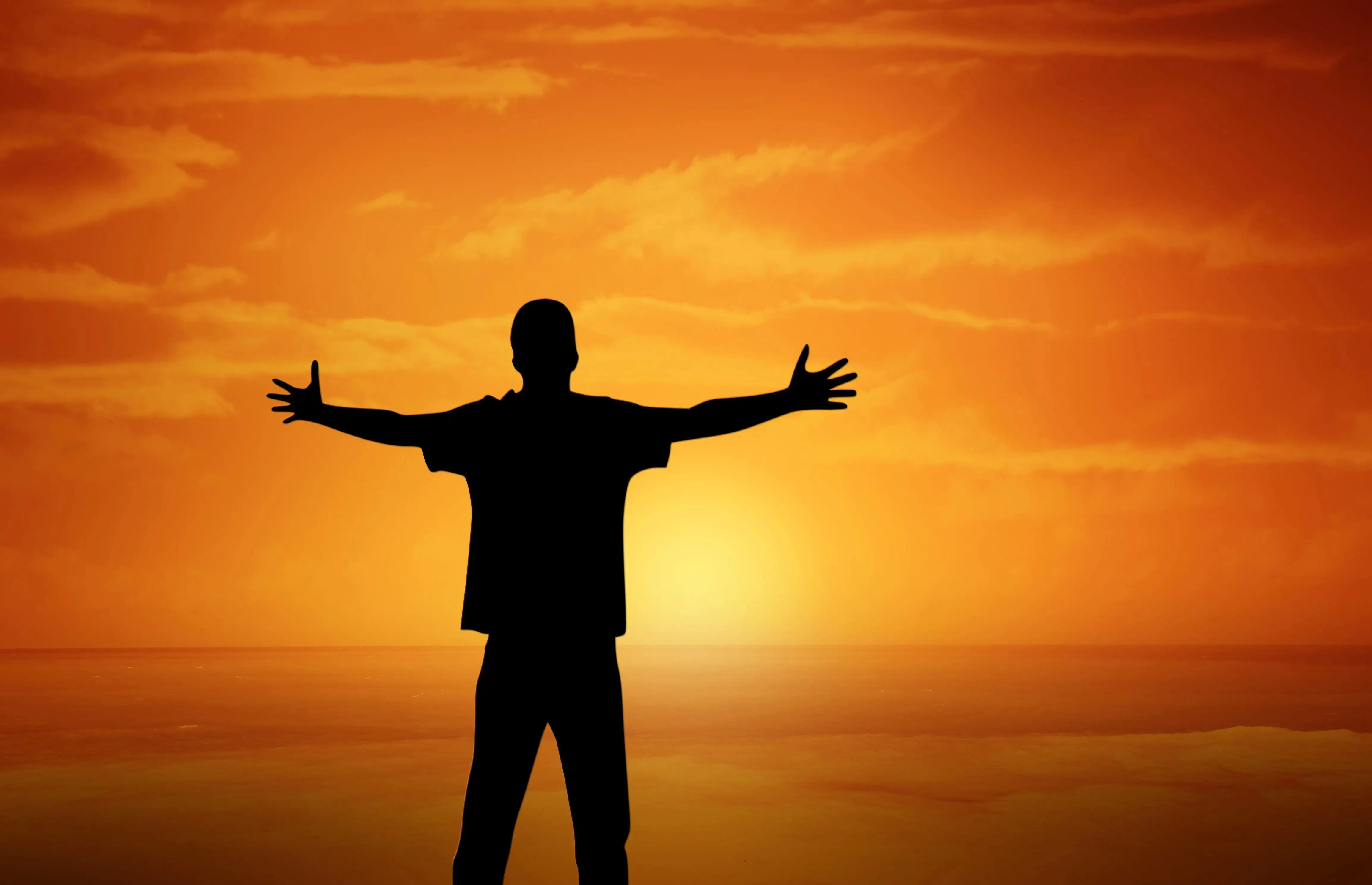 A silhouette of a person standing with arms outstretched against a vivid orange sunset sky over a calm ocean. The figure is centered in the image, facing the horizon where the sun is setting, casting a warm golden glow across the sky and water. Wispy clouds are scattered above, enhancing the dramatic and peaceful atmosphere. The image conveys a sense of freedom, gratitude, and connection with nature.